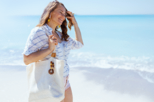 A woman with a loose fitting white button down swimsuit cover walks on the beach with her sunglasses in hand on a sunny day with the ocean in the background
